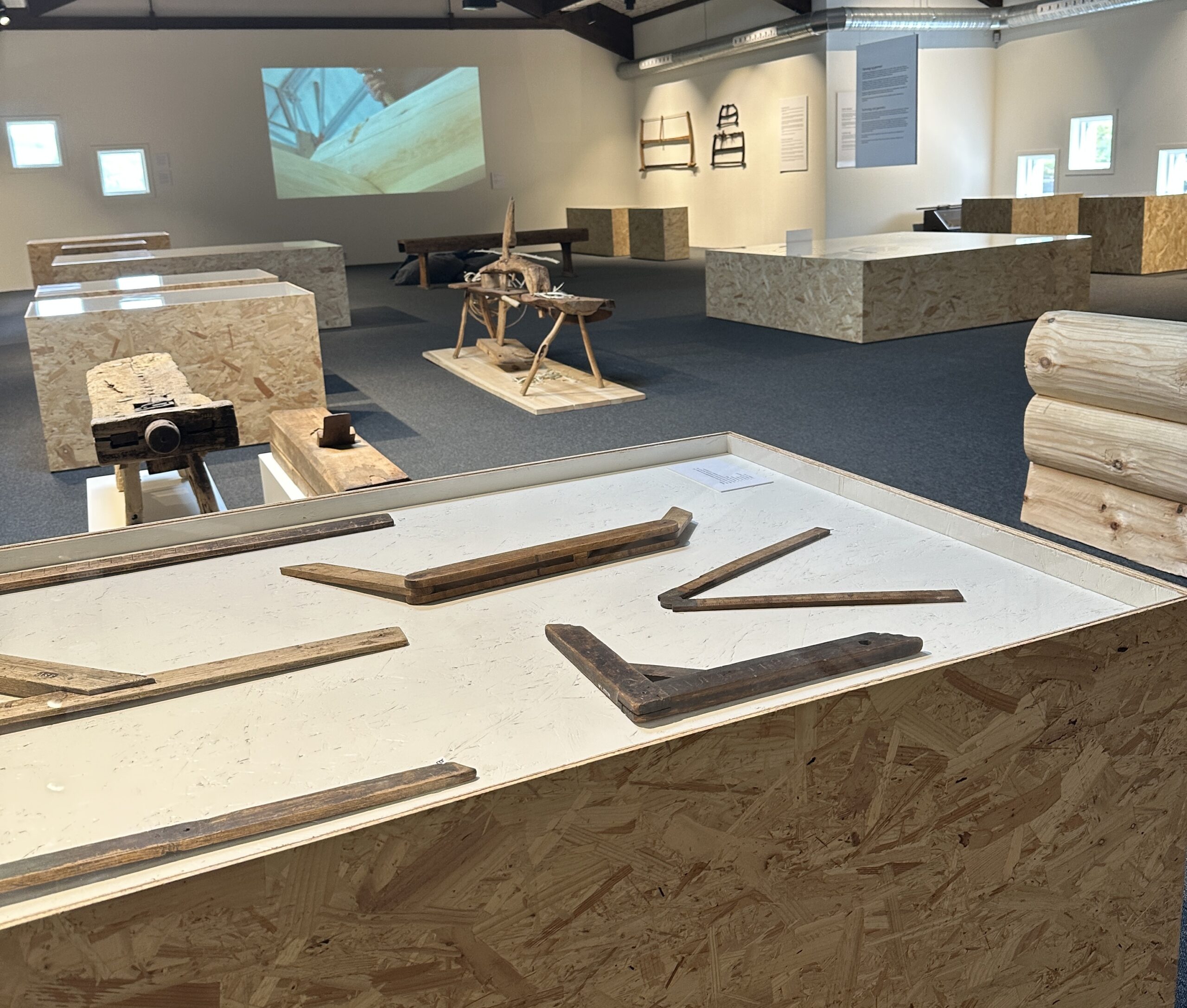 "From From Forest to Folk Art" VFK_photo_ Tine kjosås Exhibition space: White walls and dark floors, two square plywood display cases with painted interiors showing old tools. A film is projected on the wall behind the cases.