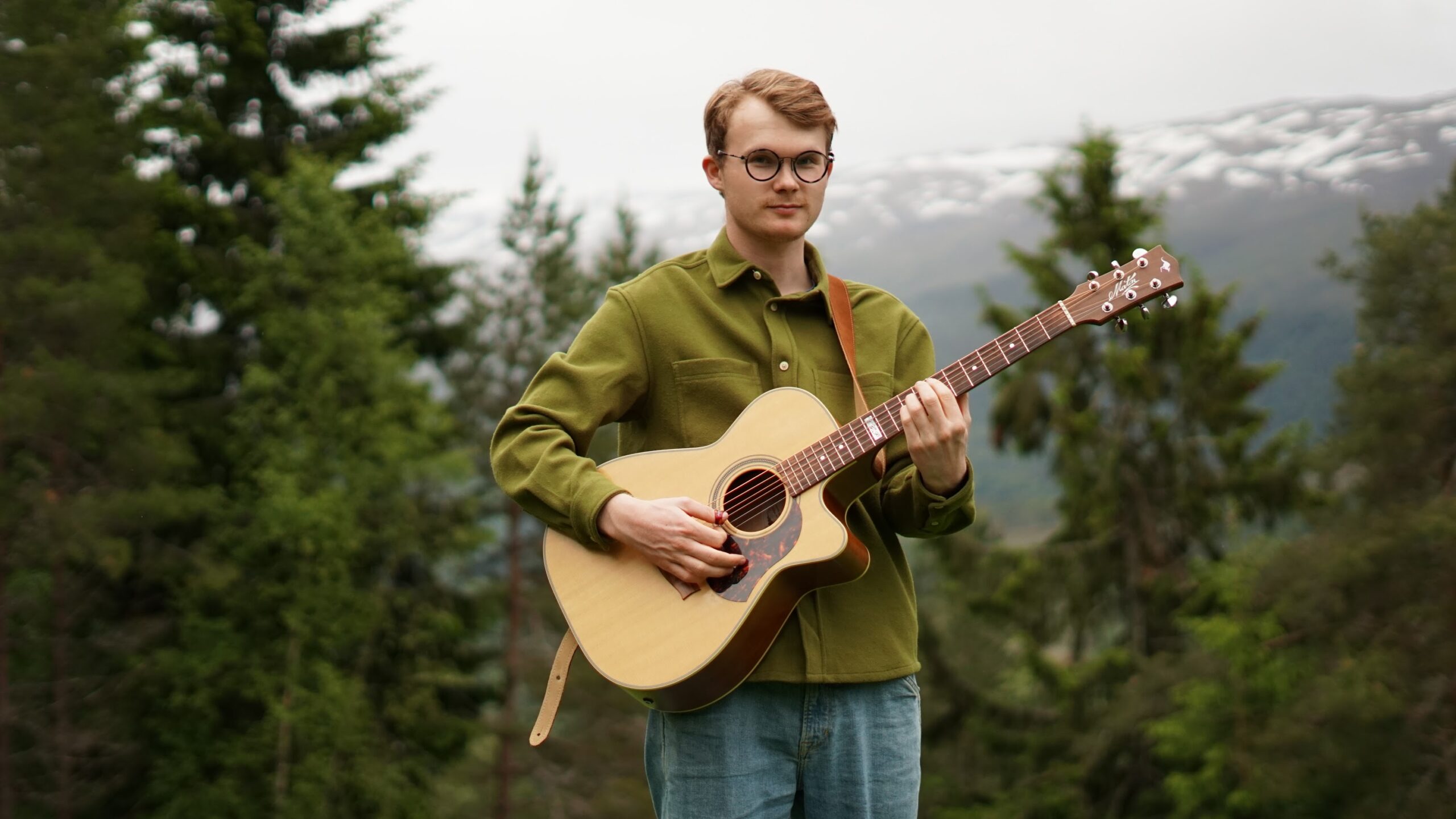 Artist picture Markus Lirhus A man wearing glasses and a green jacket stands in a spruce forest with a guitar strapped across his chest.